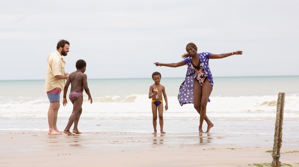 Familie links Mann in Hemd, Mitte zwei Kinder, rechts Frau in blauem Kleid, dahinter Strand.