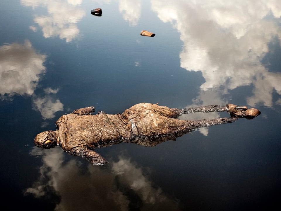 Ein Mann treibt tot im Wasser, in dem sich Wolken spiegeln.