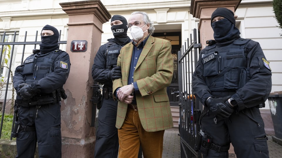 Prinz Reuss in Anzug mit Gesichtsmaske, eskortiert von drei polizeilichen Spezialeinheiten vor einem Gebäude.
