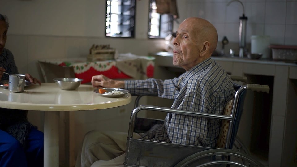 Alter Mann in einem Rollstuhl sitzt an einem Esstisch.
