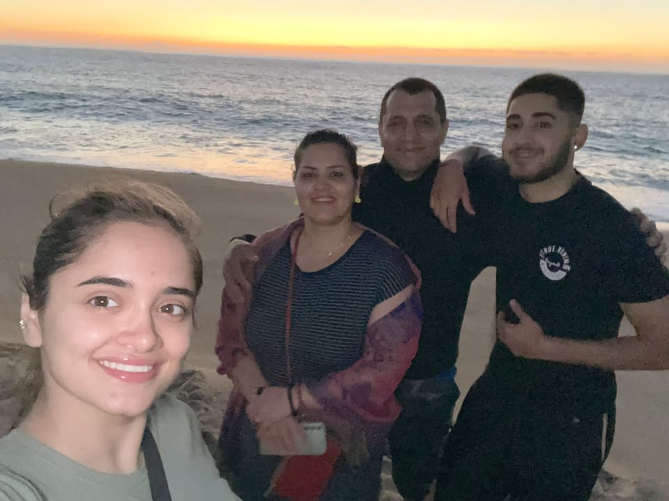Eine junge Frau steht mit ihren Eltern und ihrem Bruder an einem Strand bei Sonnenuntergang und macht ein Selfie.