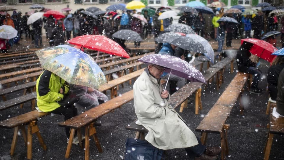 Glarnerinnen und Glarner sitzen im Ring mit aufgespannten Schirmen im Schneegestöber.