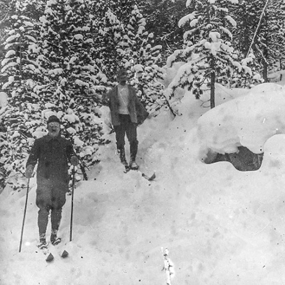 Zwei Männer mit Skiern am Waldesrand im Schnee.