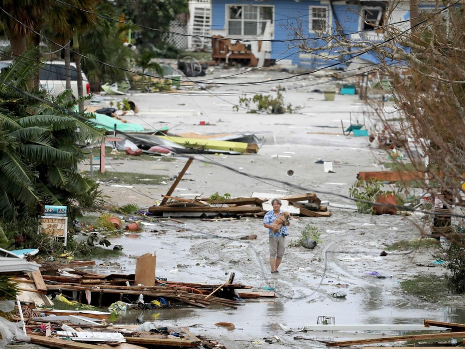 Ein Mensch trägt einen Hund, während er über eine überflutete Strasse läuft, auf der die Teile von zerstörten Häusern liegen.