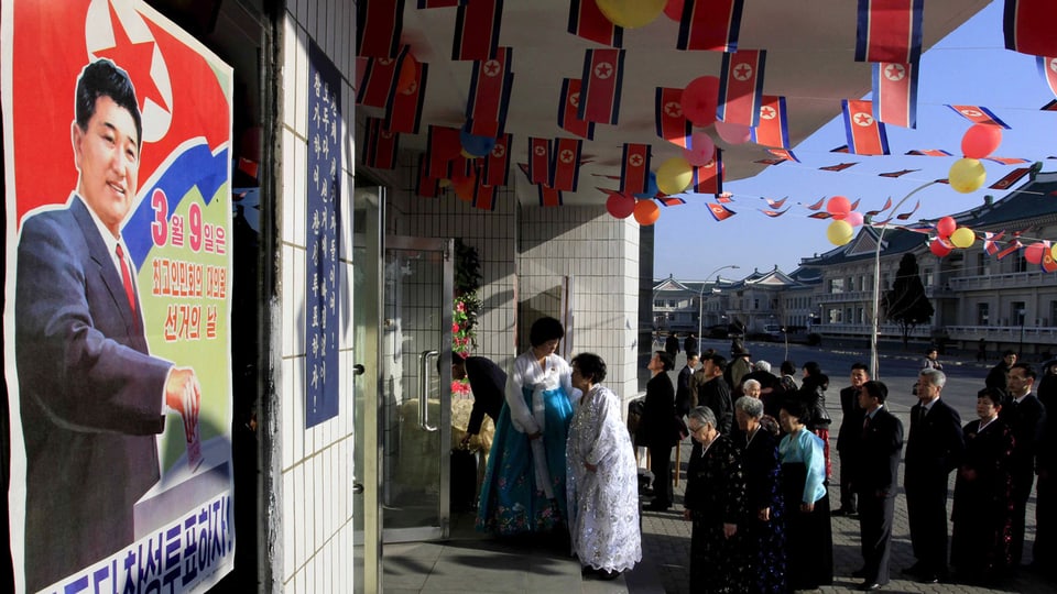 Schlange vor einem Wahllokal in Nordkorea. Kleine Fahnen schmücken die Decke der Überdachung, links ist ein Wahlplakat zu sehen