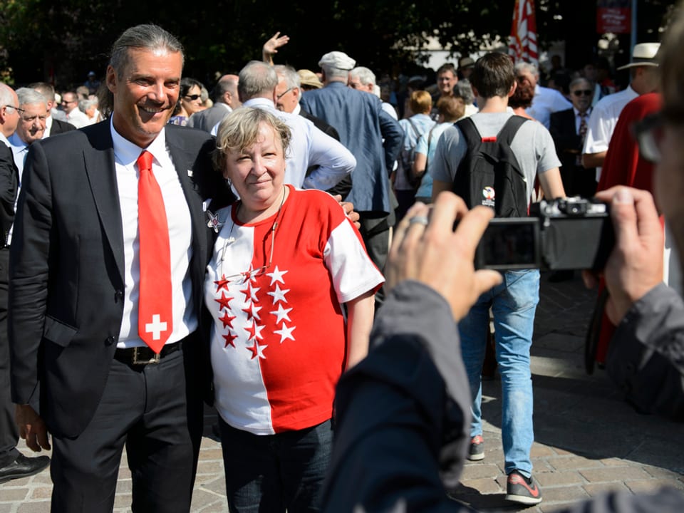 Freysinger mit Walliserin mit Walliser Flagge auf T-Shirt. Menschen im Hintergrund, Fotokamera im Vordergrund.