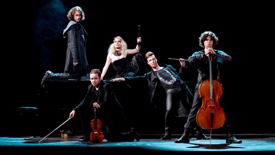 Eine Frau mit Flöte in der Mitte, um sie herum schwarzgekleidet und düster blickend 4 weitere Musiker.
