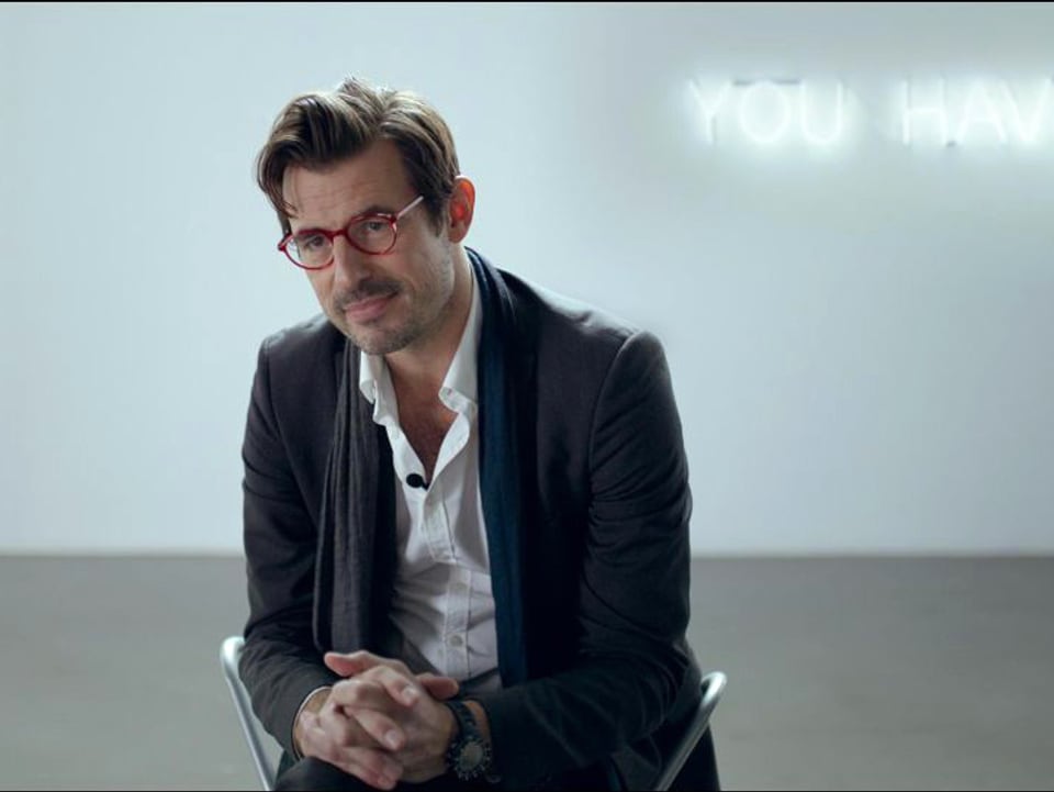 Ein Mann mit chnauz und Brille sitzt in einem leeren Raum. 