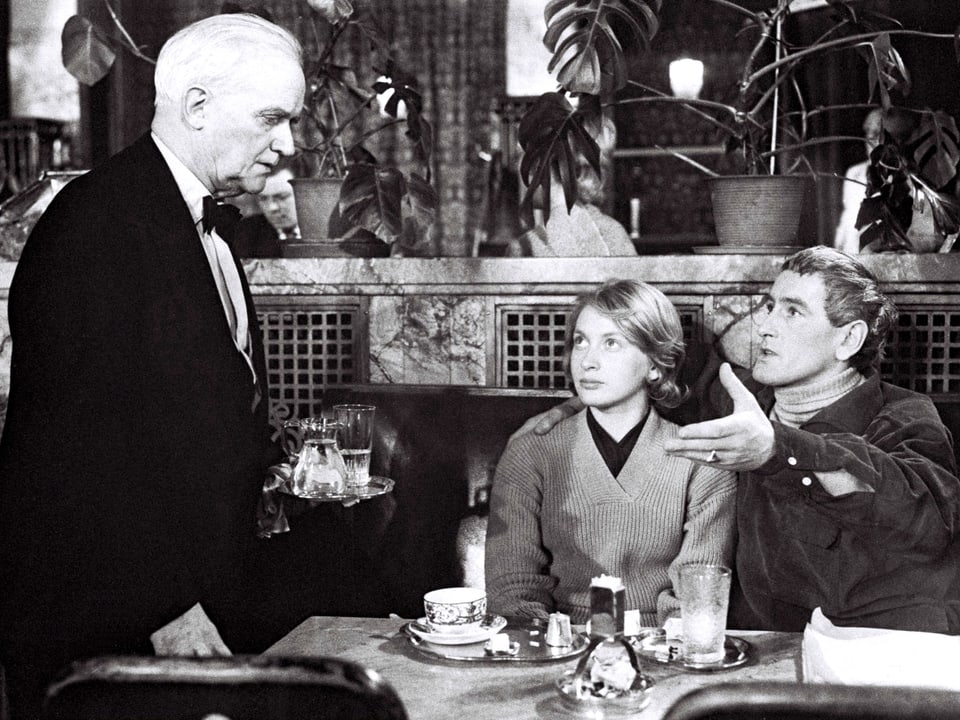 Ein junges Paar sitzt an einem Tisch in einem Café vor sitzen Getränken und sprechen mit einem älteren Kellner.
