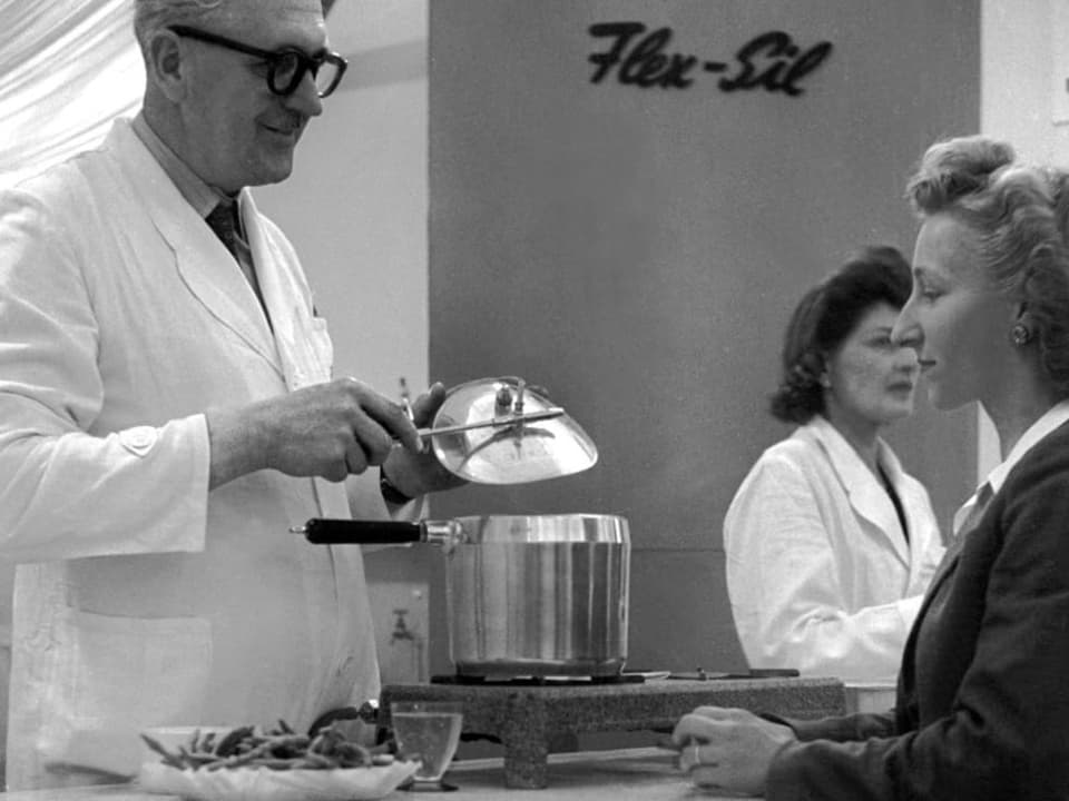 schwarz-weiss Foto: Rechts eine Frau, schaut nach links in einen Dampfkochtopf, gezeigt von Mann in weissem Kittel.