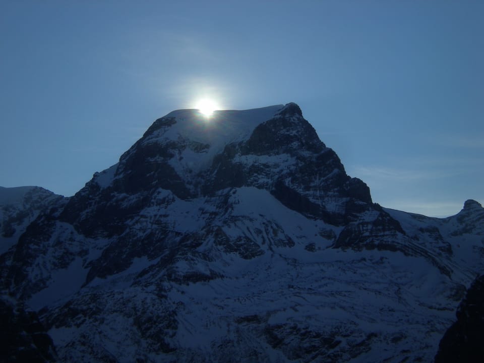 Der Himmel ist blau. Unten sieht man ein Bergmassiv mit etwas Schnee. Die Sonne ist knapp über einer Bergkante zu erkennen.