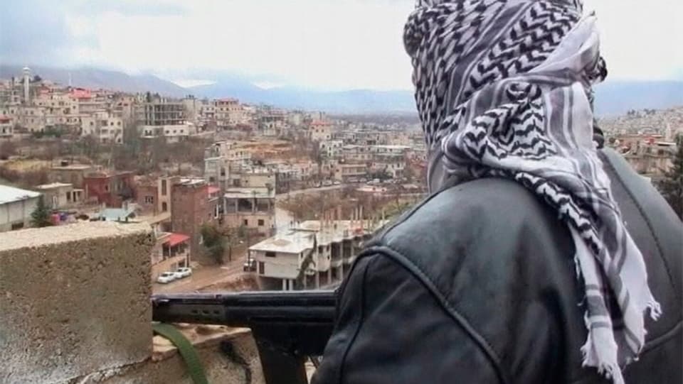 Rückenansicht eines Mann mit Kopftuch und Gewehr, der von einer Mauer auf den Ort Zabadani schaut.