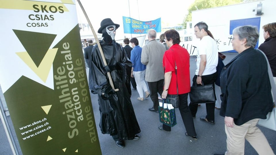 Ein als Tod verkleideter Demonstrant steht neben einem Plakat der Skos.