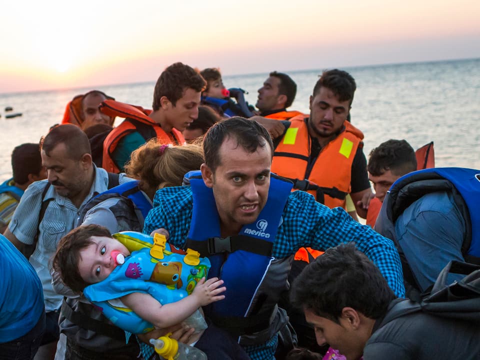 Ein Mann trägt ein etwa einjähriges Kleinkind auf dem Arm. Hinter ihm viele andere Flüchtlinge. 