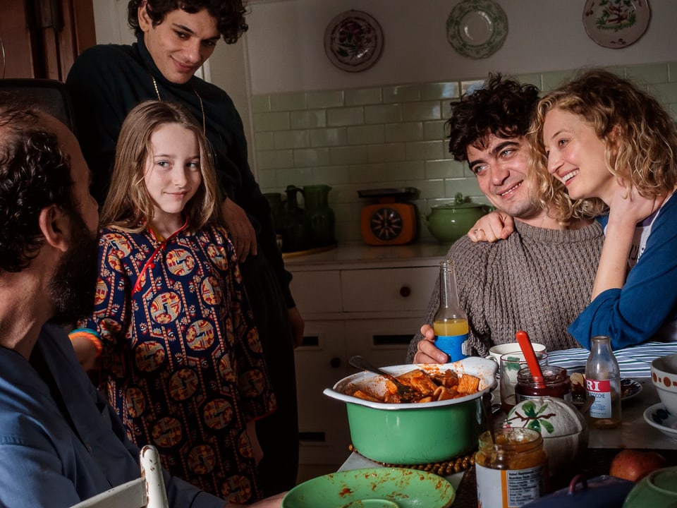 Filmszene: Eine Familie (älterer Mann, zwei junge Männer, junge Frau und ein Mädchen) sitzen lachend an einem Küchentisch