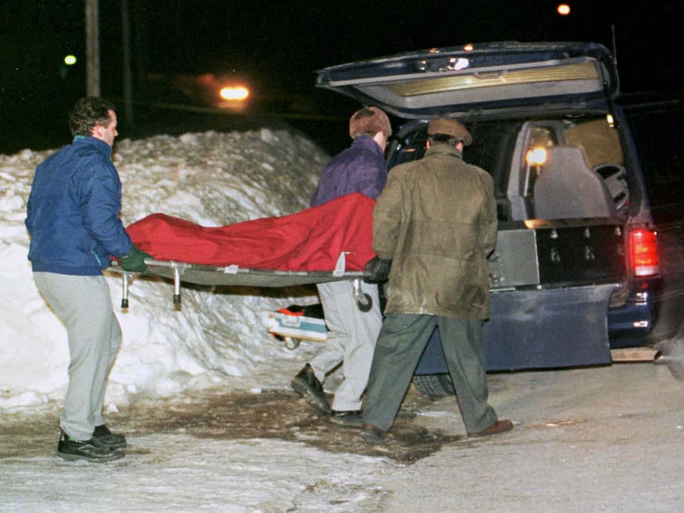 Drei Männer tragen eine Bahre zu einem Auto, auf dem ein Toter liegt. Er ist von einem roten Tuch bedeckt.
