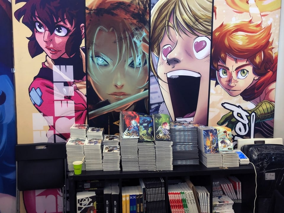 Auf einem Tisch liegen Mangas in Stapeln auf. Im Hintergrund sind grosse Werbeplakate von Manga-Büchern.