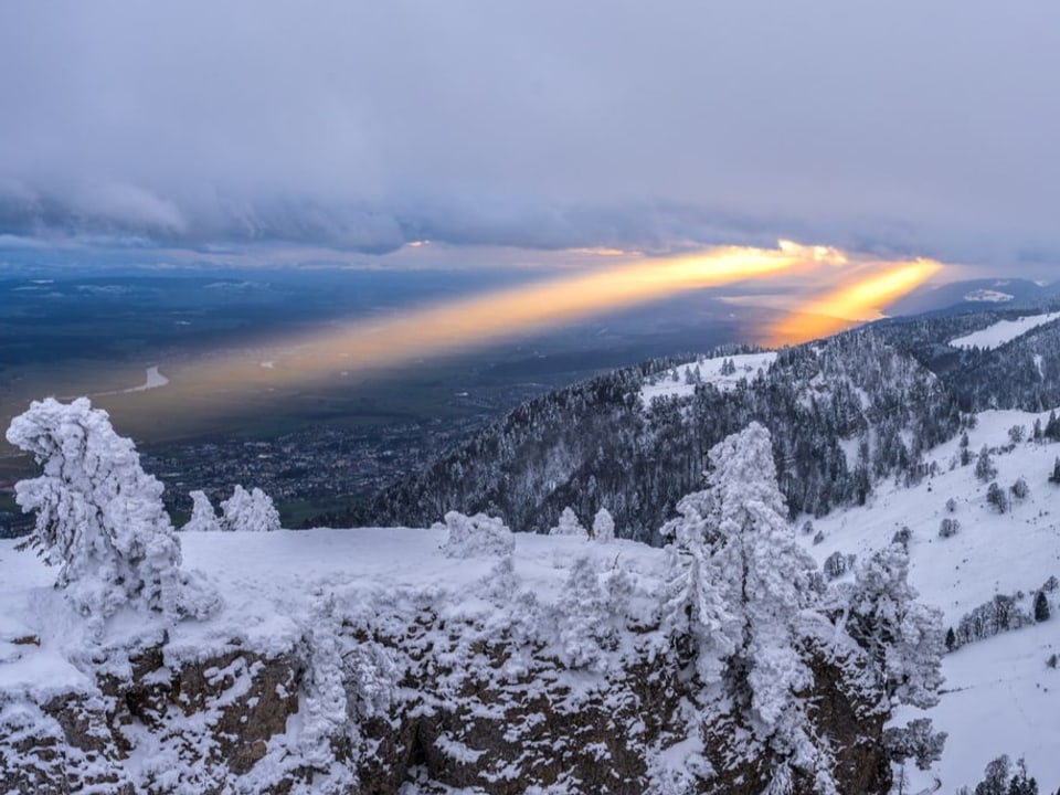 Karger Hügel mit Schnee und Eis, Himmel grau. Sonne drückt wie zwei Scheinwerfer durch die Wolken. 