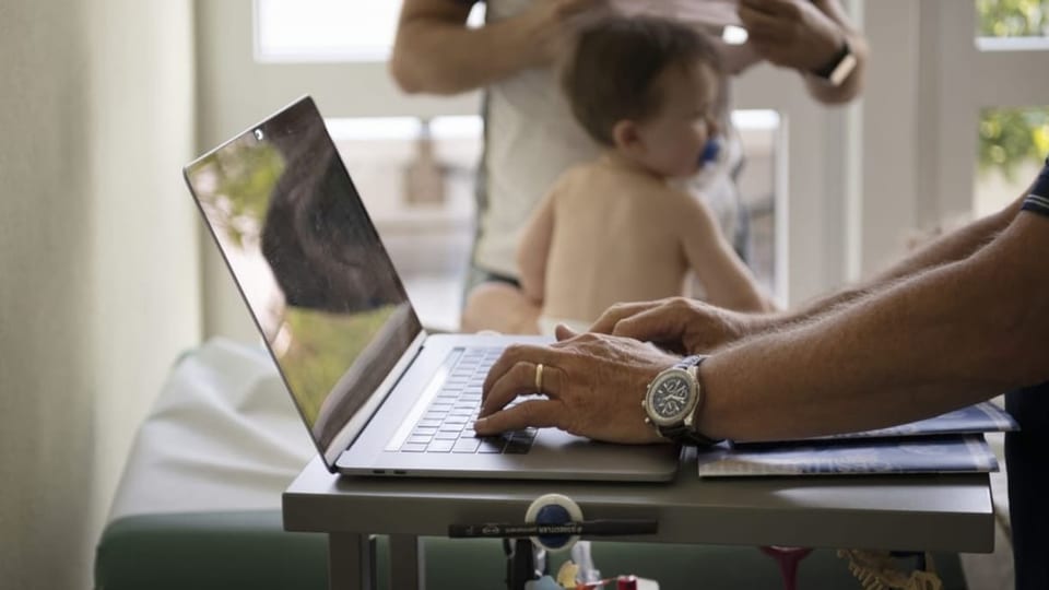 Arzt an Laptop – im Hintergrund sieht man ein Kleinkind