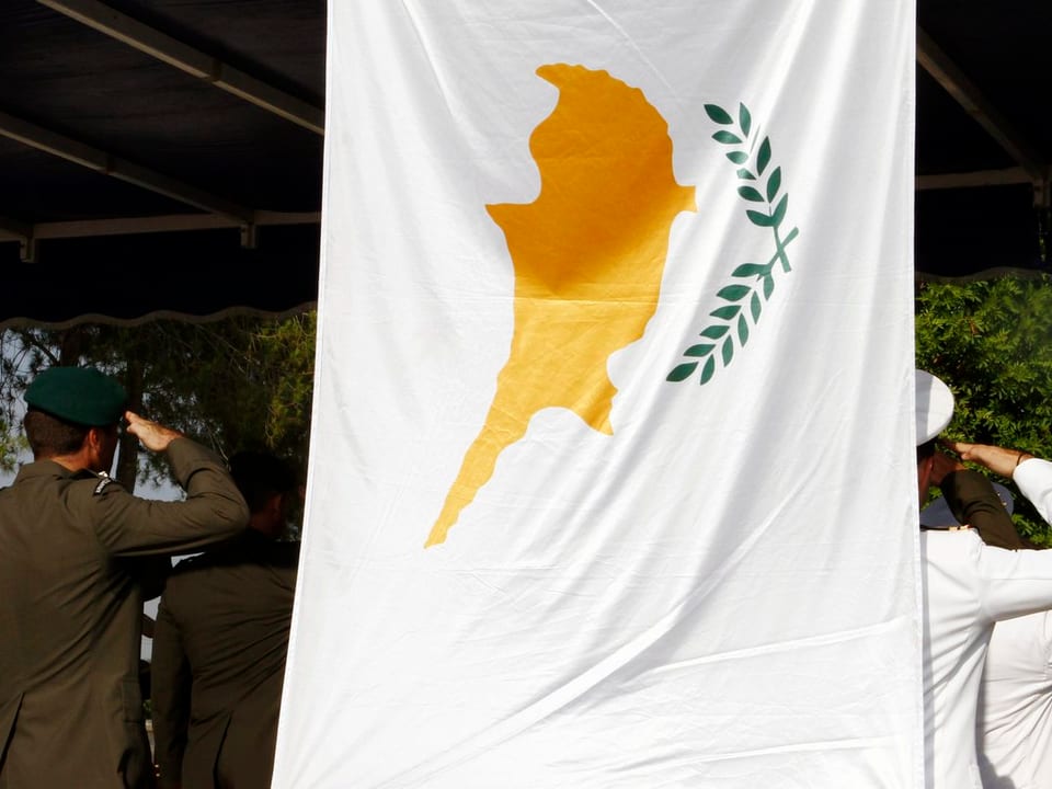 Soldaten salutieren. Sie stehen unter einem Dach. Im Vordergrund weht die zypriotische Flagge