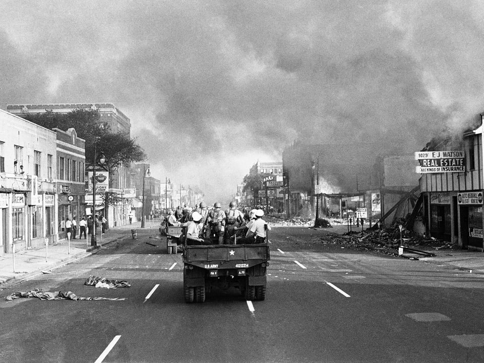 Brennende Gebäude und Qualm. Im Vordergrund Militär auf einem Jeep sitzend. (Schwarz-Weiss)