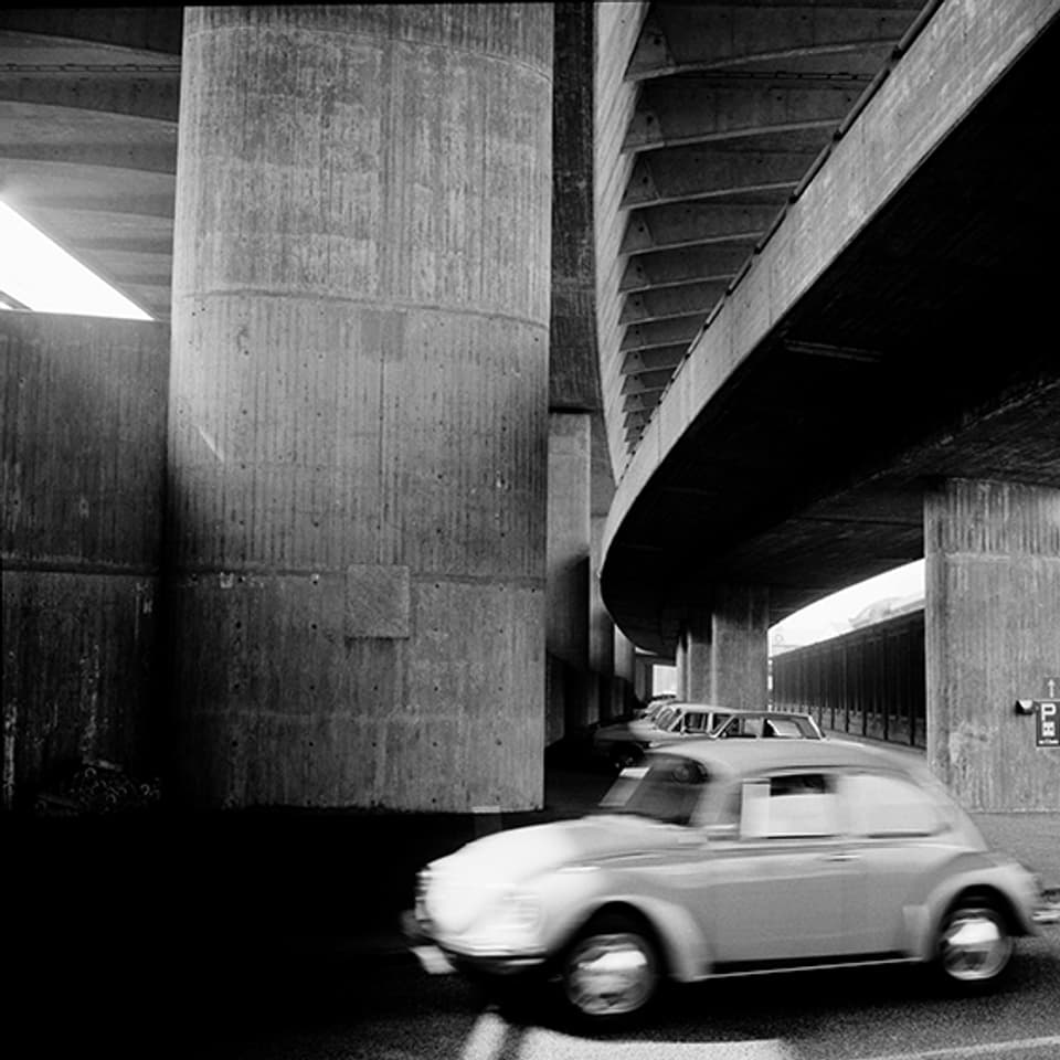 Auf einer schwarz-weissen Fotografie ist die Unteransicht einer Beton-Brücke zu sehen, ein Auto fährt auf einer Strasse vorbei.