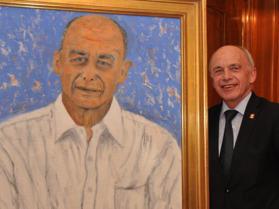 Ein Mann lächtelt in die Kamera, links neben ihm ein Gemälde von ihm.