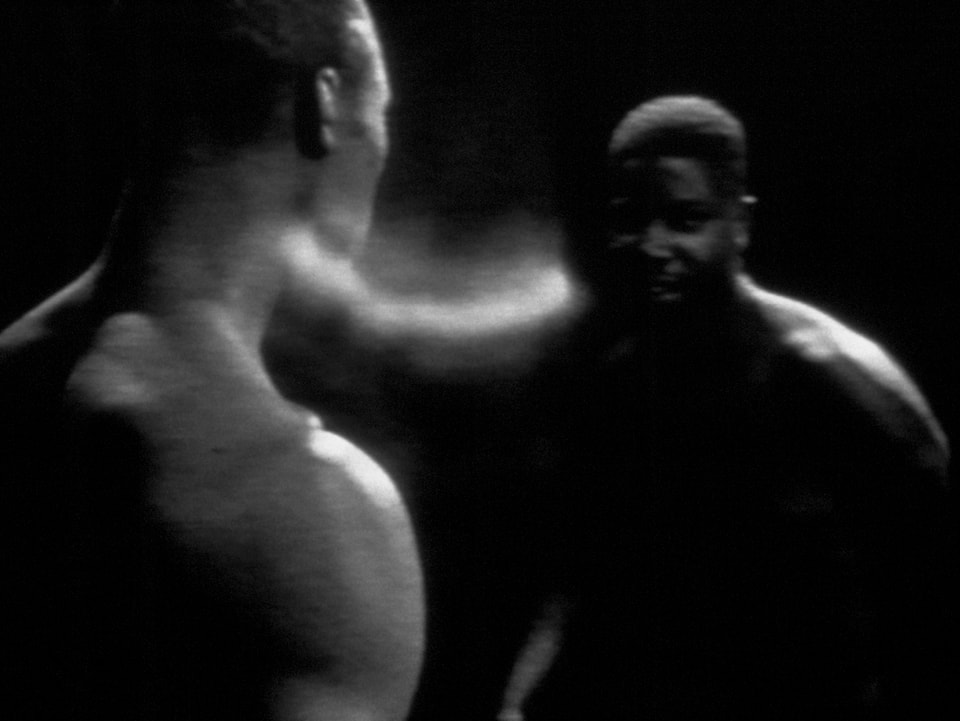 Schwarz-weiss Aufnahme von Steve McQueen, auf dem im Dunklen zwei dinkelhäutige Körper zu sehen sind, die durch einen Lichtschein verbunden zu sein scheinen.
