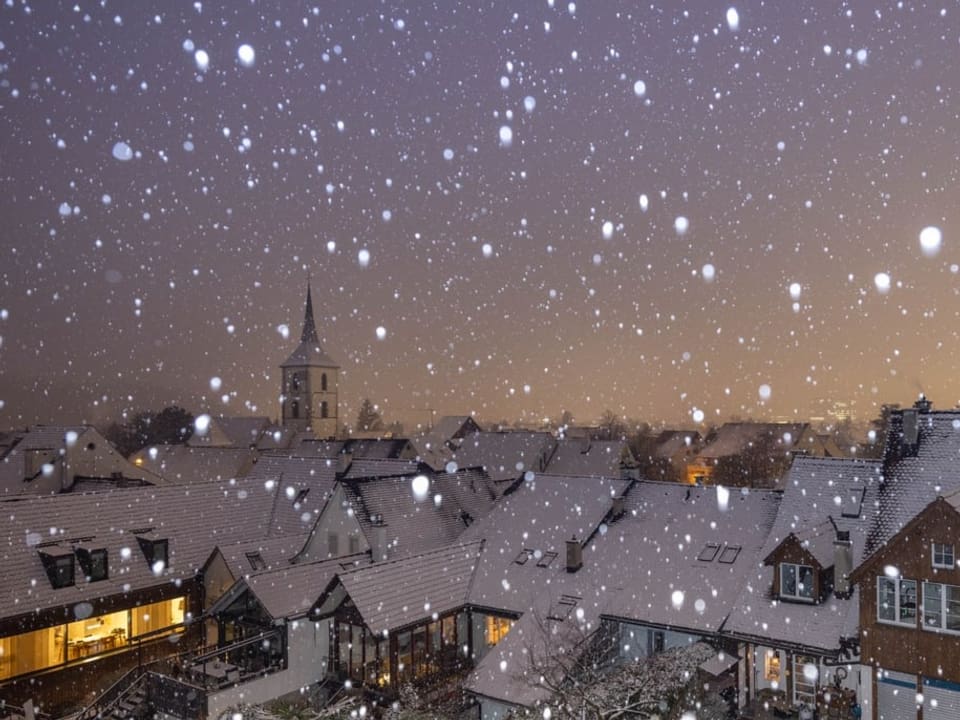 Dorf mit verschneiten Häusern, auch durch die Luft tanzen weitere Schneeflocken in der Morgendämmerung.