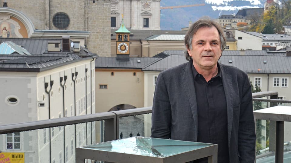 Ein Mann mit grau meliertem, halblangem Harr steht auf einer Terrasse. Im Hintergrund ist eine Kirchenanlage zu sehen.