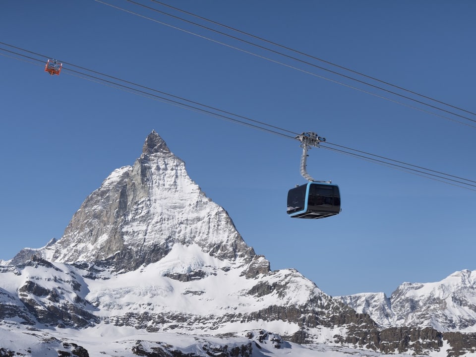 Eine Seilbahnkabine fährt vor einem verschneiten Bergpanorama durch.