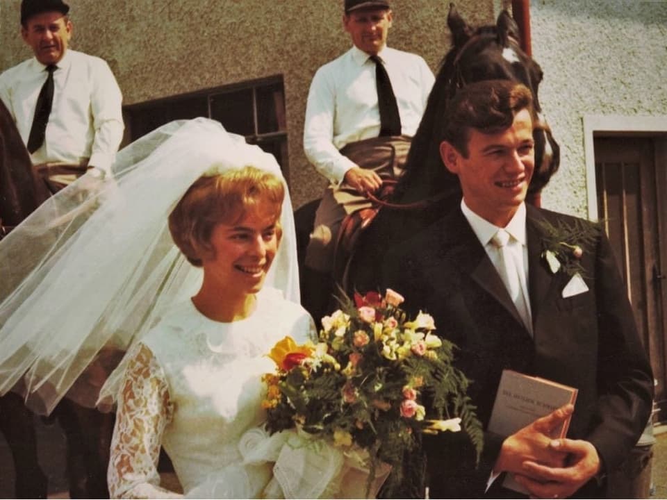 Ein junges Ehepaar steht lächelnd vor zwei Pferden mit Reitern. Die Braut trägt einen grossen, farbigen Blumenstrauss. Ihr Schleier weht im Wind.