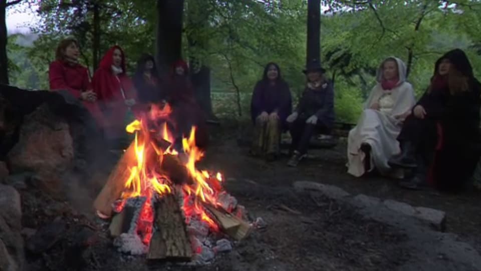 Feuer im Wald, dahinter sitzt eine Runde von Frauen in farbigen Umhängen