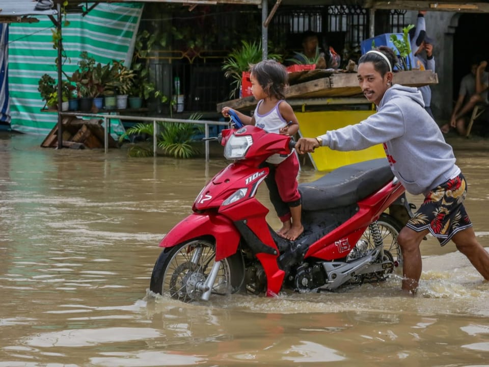 Ein Mann schiebt sein Motorrad mit einem Kind an Bord durch eine überflutete Strasse auf den Philippinen.