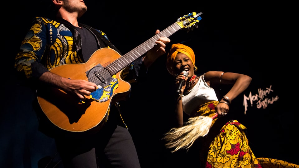 Ein Mann mit Gitarre und eine Frau in afrikanischer Tracht auf der Bühne.