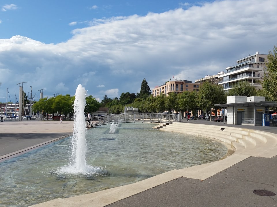 Der Himmel wird durch eine Wolkenwand auf der rechten Seite zweigeteilt. Im Vordergrund steht der Springbrunnen von Lausanne Ouchy.