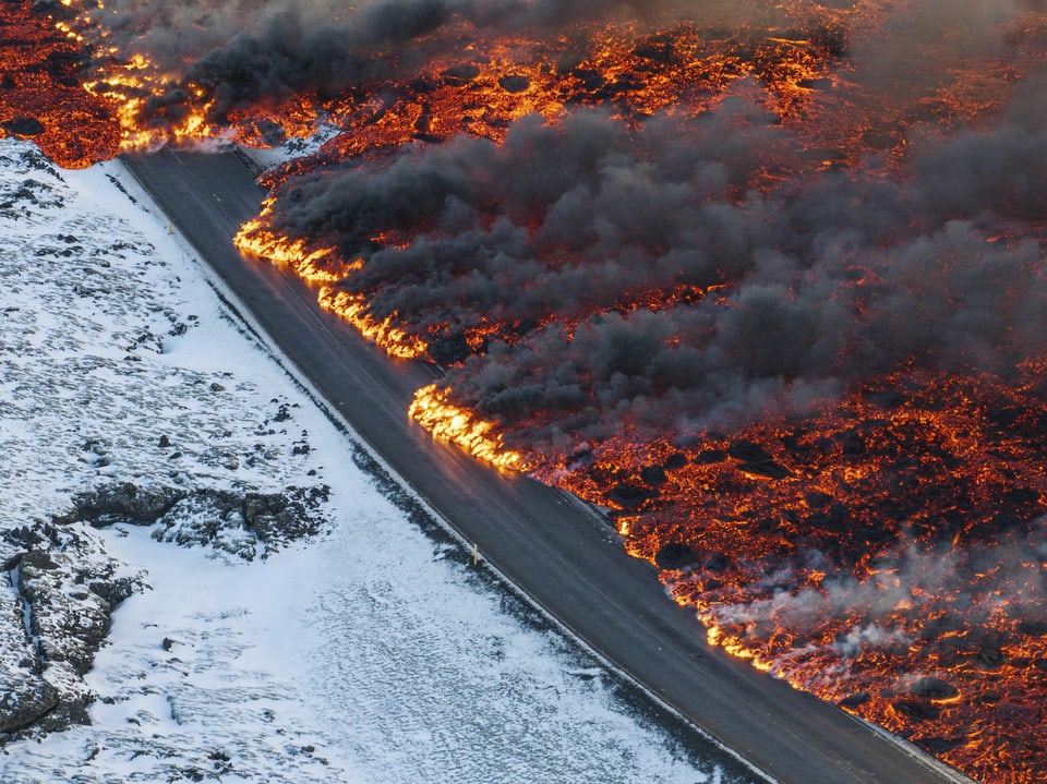 Rotglühende Lava fliesst auf die alphaltierte Strasse. Die Umgebung ist schneebedeckt.