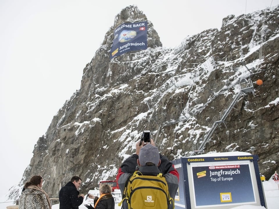 Touristen fotografieren eine Fahne am Gipfel des Berges: Welcome Back - heisst es da drauf.