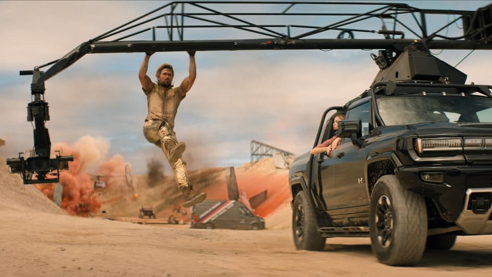 Mann hängt an Kameraausleger über einem Filmset mit Explosion und Truck in der Wüste.