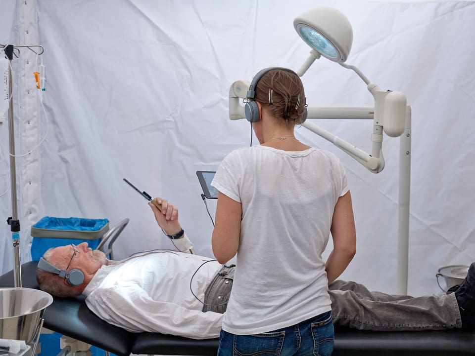 Ein Mann und eine Frau in einem Operationssaal, beide mit Kopfhörern und tablets. Er liegt auf der Liege, sie steht daneben.