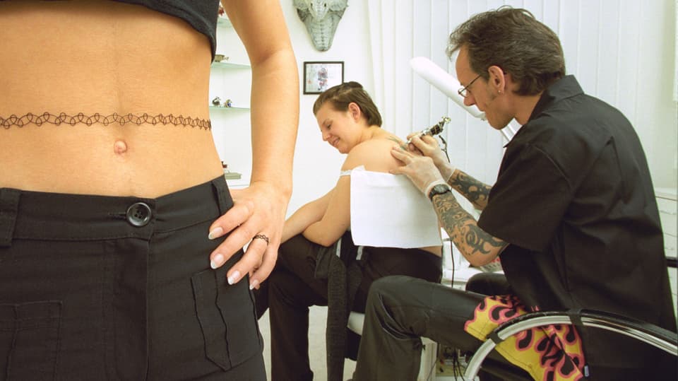 Links ein nackter Bauch, darum ein schwarzes Tattoo-Stretch-Bändchen, rechts wird eine Frau tätowiert von einem Mann.