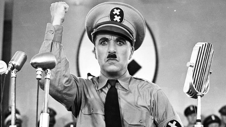 Charlie Chaplin, verkleidet als Adolf Hitler, steht vor Mikrofonen und streckt mit strenger Mine die Faust in die Luft.