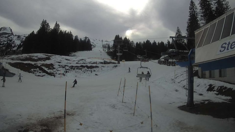 Sesselbahn und Skifahrer auf weisser Piste, grüne Flecken