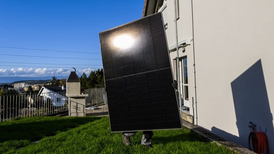 Ein Monteur trägt ein grosses Solarpanel zu einem Wohnhaus.