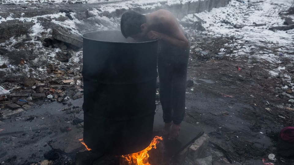 Junger Mann mit nacktem Oberkörper und barfuss wäscht sich draussen in der Kälte mit warmen Wasser aus einer Tonne, die über dem offenen Feuer erhitzt wird
