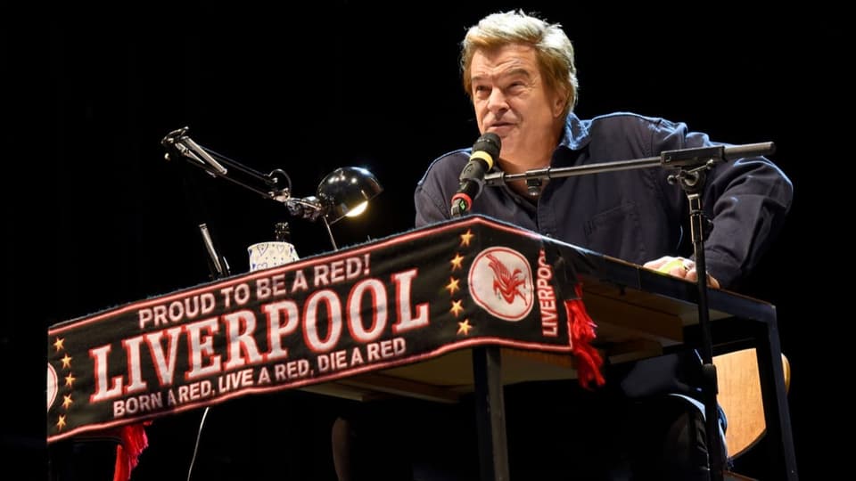 Mann an Rednerpult mit Mikrofon, an Pult hängt Banner von FC Liverpool.