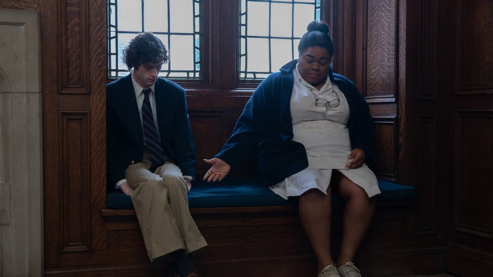 Ein junger weisser Mann und eine mittelalte schwarze Frau sitzen auf einer Bank. Sie streckt ihre Hand nach seiner aus.