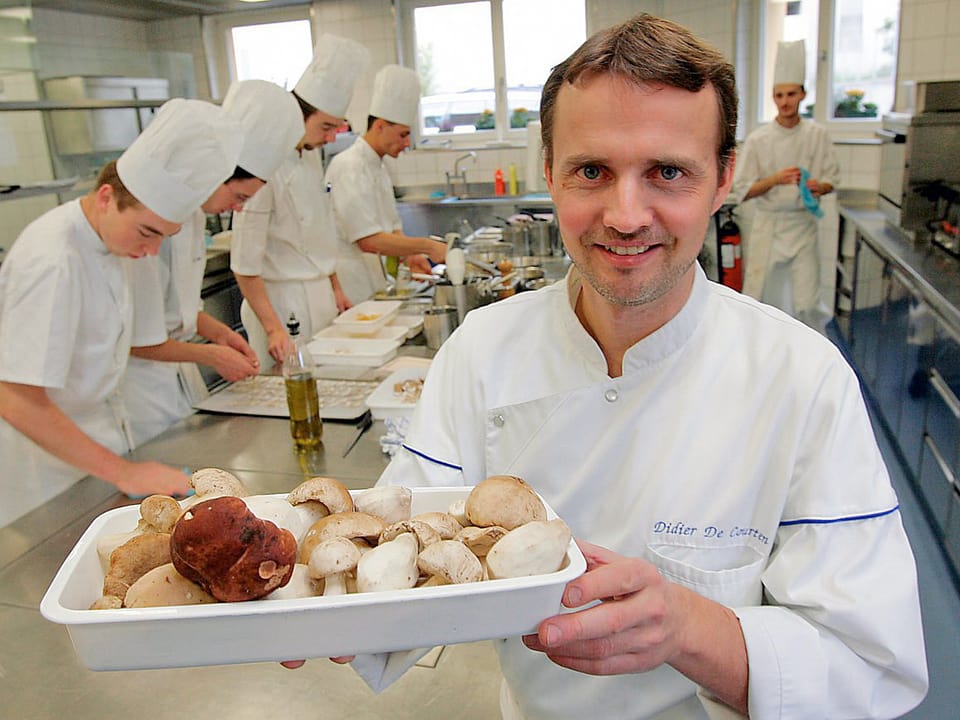 Didier De Courten mit einer Schale mit frischen Pilzen, im Hintergrund sind vier Köche am Werk.