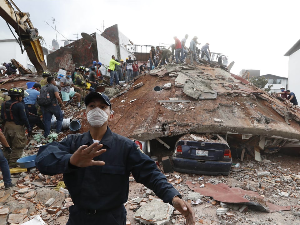 Mann mit Staubmaske im Vordergrund, dahinter ein zusammengebrochenes Gebäude.