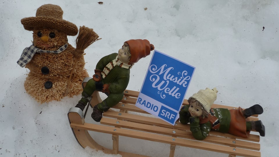 Ein Schneemann aus Stroh und zwei Puppen, ein Bube und ein Mädchen. Sie sitzen auf einem Holzschlitten, dazwischen steht das Logo von SRF Musikwelle.